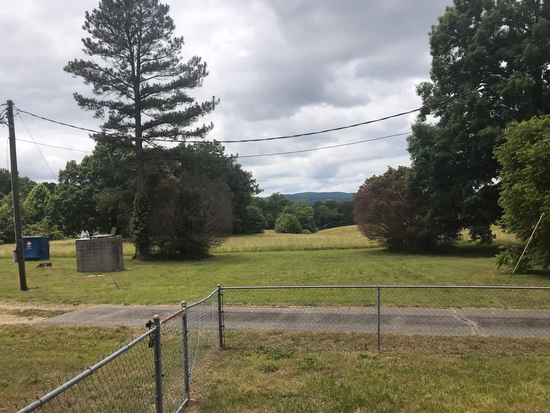 A grassy outdoor area with a chain-link fence in the foreground, a paved path, several trees, a utility pole with power lines, a blue dumpster, and a small concrete structure under a cloudy sky.