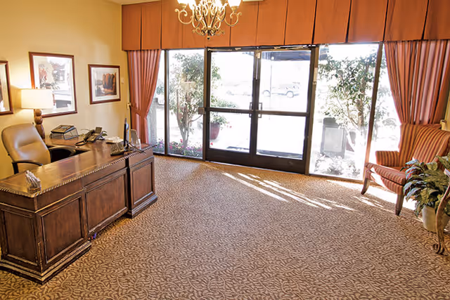 A well-lit office area with a large wooden desk and a leather office chair. The desk has a telephone, a computer monitor, and some office supplies. There are framed pictures on the wall, a chandelier hanging from the ceiling, and large glass doors with curtains leading outside. A striped armchair and a potted plant are positioned near the glass doors.