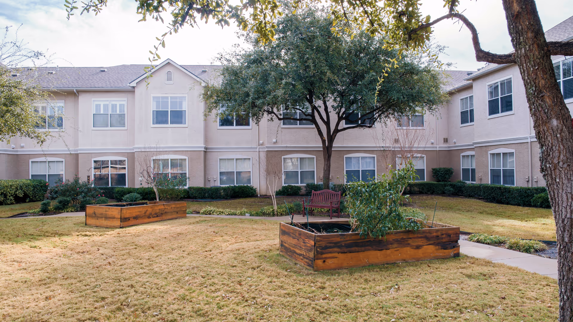 Courtyard with raised wooden planters, a bench and trees in front of a two-story beige senior living building.