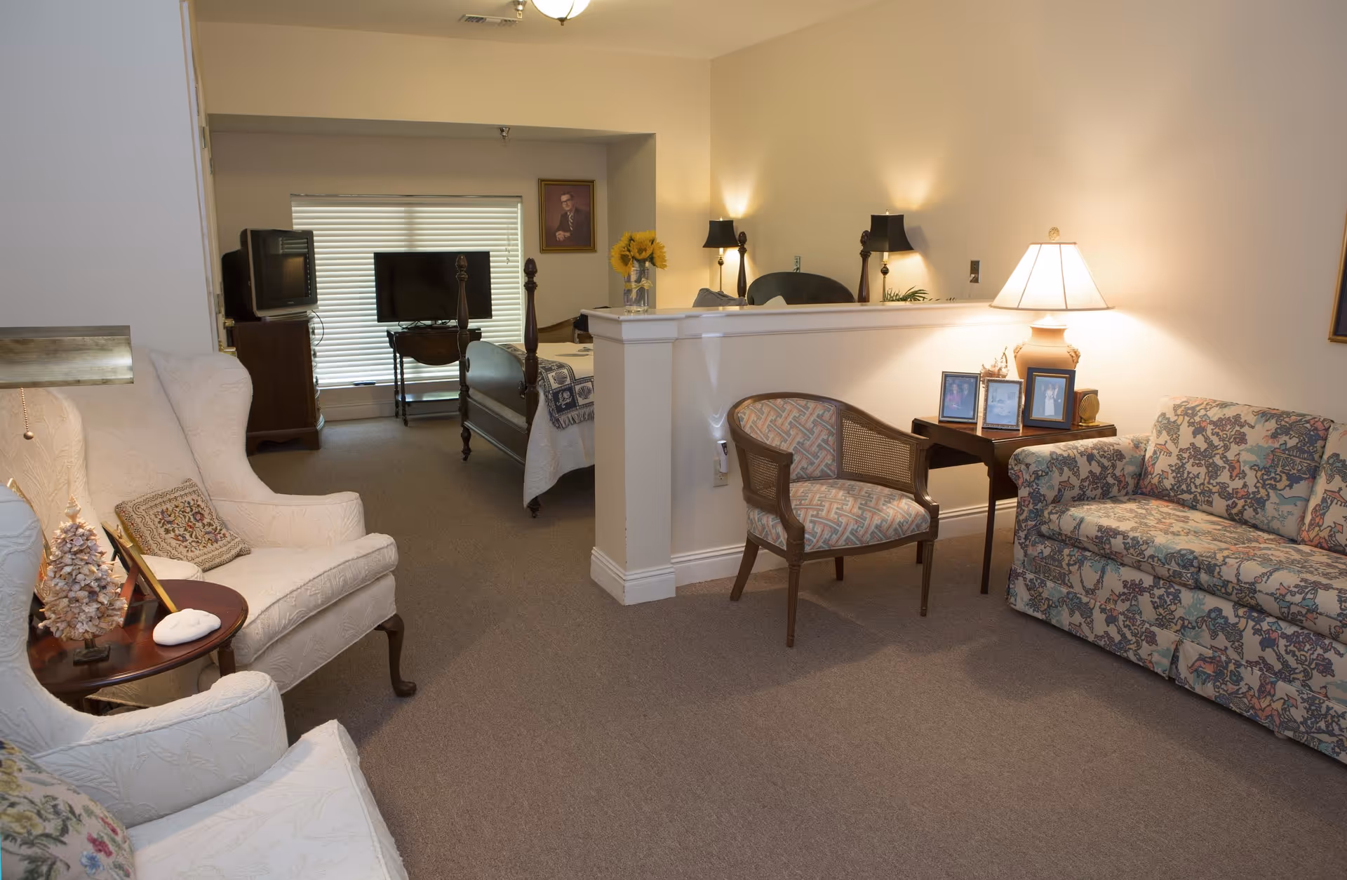 A cozy living area with a floral-patterned sofa, an upholstered armchair, a wooden chair with patterned fabric, and a small table with framed photos and a lamp. Behind a half wall, a bedroom is visible with a wooden bed, two televisions, a window with blinds, and a portrait on the wall.