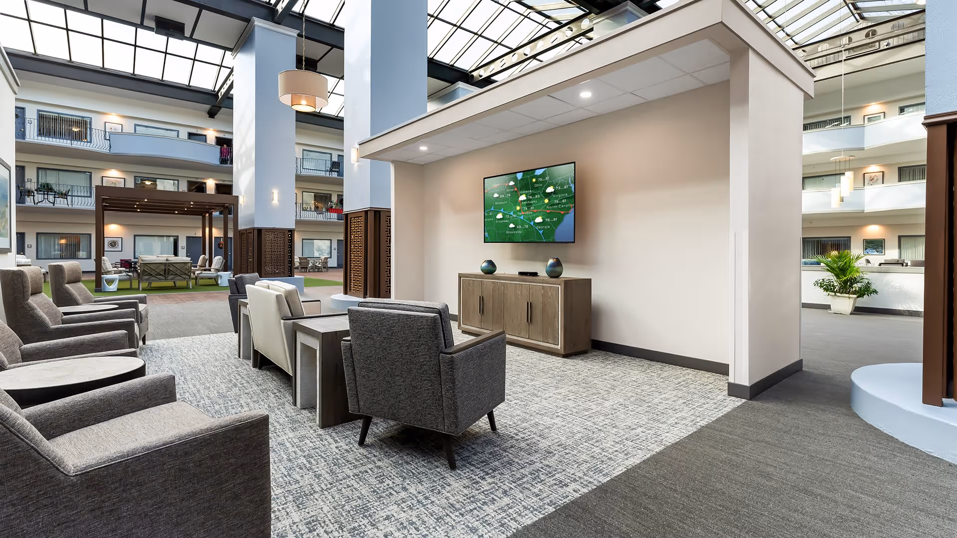 A spacious indoor common area with multiple comfortable armchairs arranged around a TV mounted on a wall. The area has a high ceiling with large skylights allowing natural light to fill the space. The background shows multiple floors with railings and doors, and some potted plants are visible.