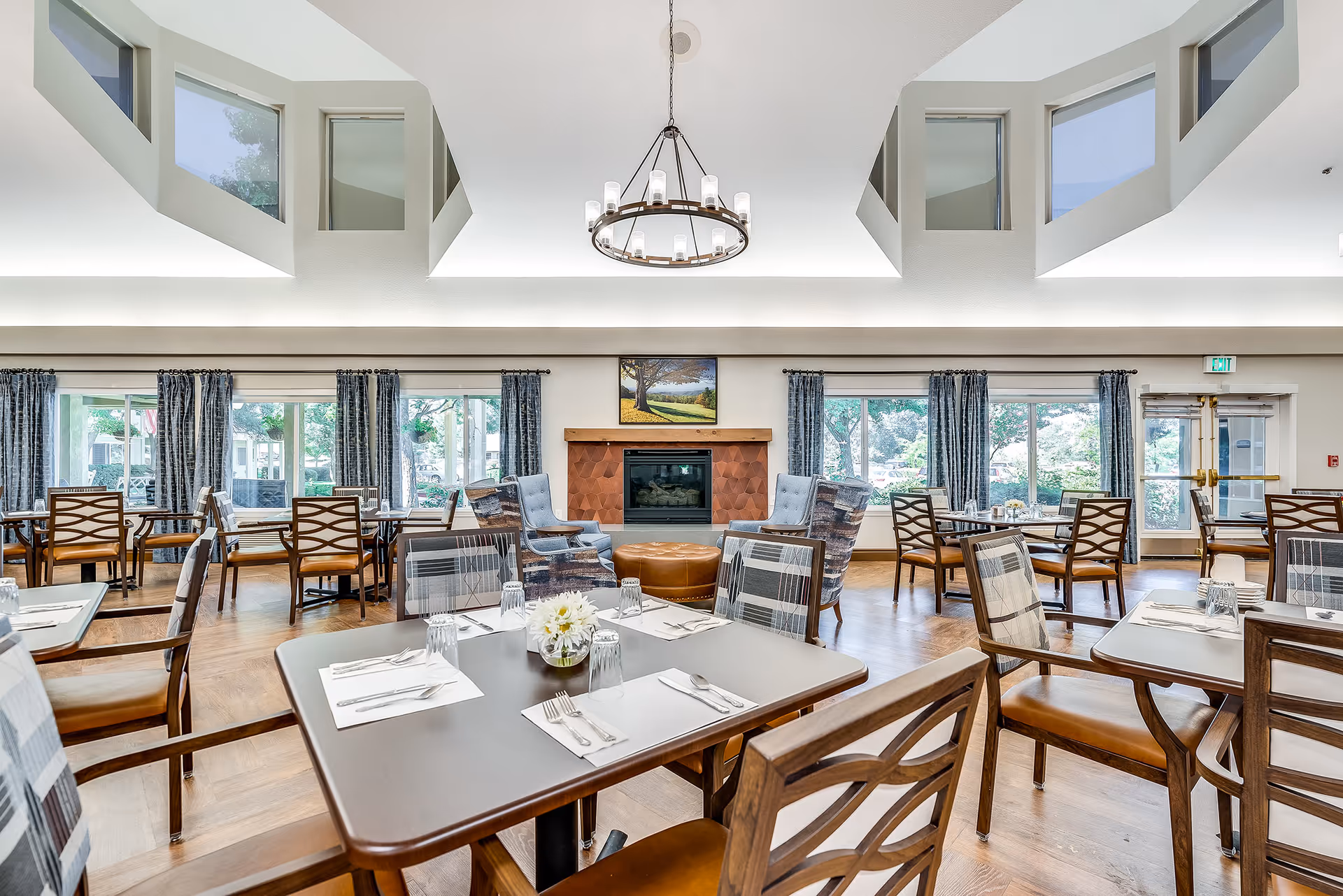 Bright dining room with multiple set tables and chairs, a central fireplace, large windows, and a high ceiling with a chandelier.