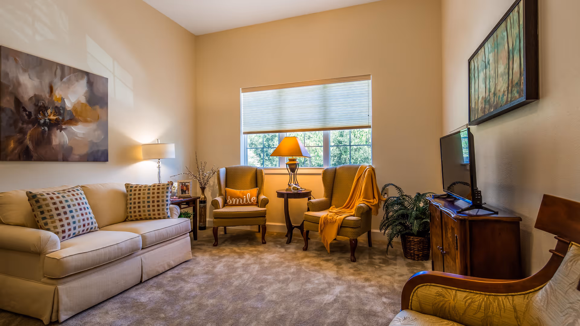 A cozy living room with a beige sofa adorned with patterned pillows, two armchairs with one draped in a yellow blanket and a 'smile' pillow, a wooden side table with a lamp between the chairs, a TV on a wooden cabinet, a potted plant, and two paintings on the walls. A window with a partially closed blind lets in natural light.