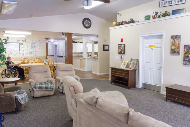A cozy common area in a senior living facility with beige upholstered chairs and a sofa arranged on a carpeted floor. In the background, there is a dining area with wooden tables and chairs, where two elderly individuals are seated, one in a wheelchair. The walls are decorated with artwork and a sign that reads 'COUNTRY LIVING'. The space has a warm, inviting atmosphere with a ceiling fan and soft lighting.
