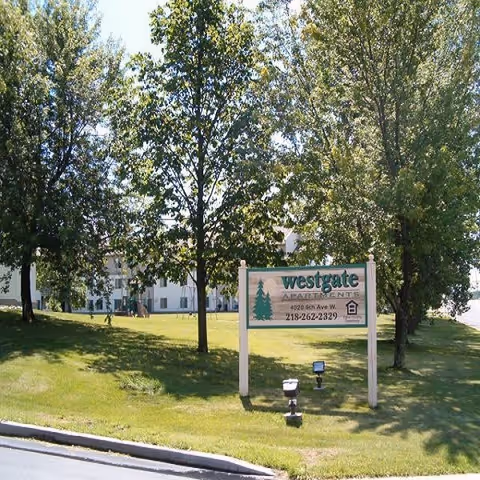 Outdoor view of Westgate Apartments with a sign displaying the name, address, and phone number. The sign is surrounded by green grass and several tall trees, with the apartment building partially visible in the background.
