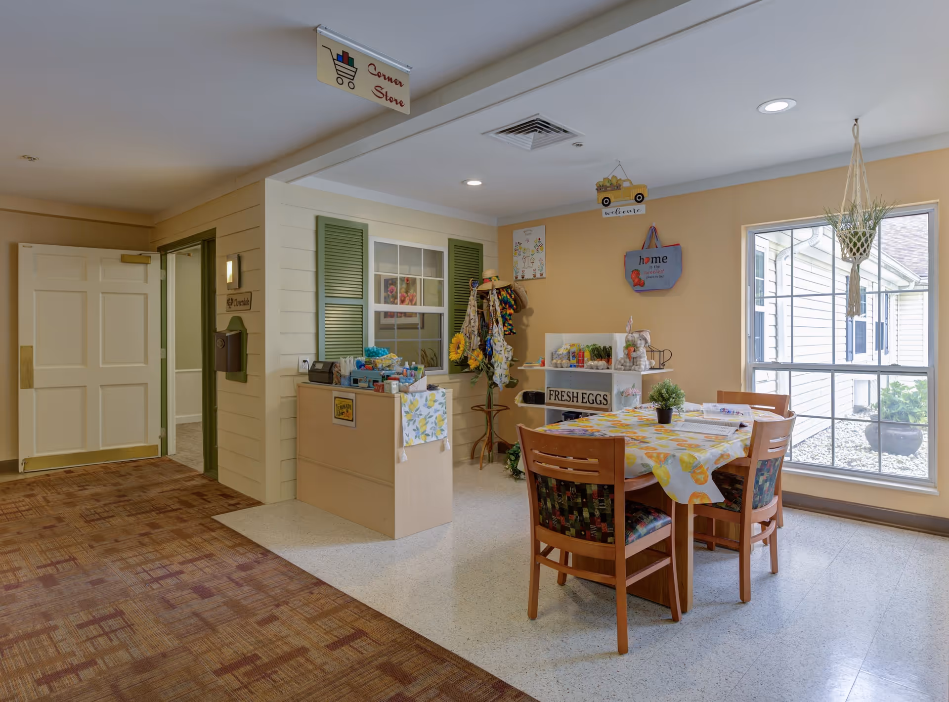 A cozy corner store area inside a senior living facility with a small table covered with a lemon-patterned tablecloth and four wooden chairs. There is a display shelf labeled 'FRESH EGGS' with various items, a coat rack with hats and bags, and a window letting in natural light. The walls are painted beige, and there is a sign hanging from the ceiling that reads 'Corner Store.'