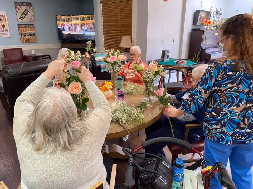 A group of elderly women sitting around a round wooden table arranging flowers in vases inside a cozy room. One woman is standing and assisting another. The room has comfortable chairs, a piano, a television, and decorative wall art.