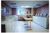 A laundry room with multiple white washing machines and dryers lined up against two walls. There is a folding table in the center of the room and a window at the far end letting in natural light. The walls are painted a muted purple color.