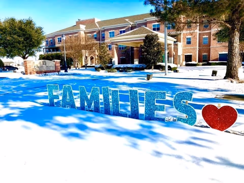 Snow-covered lawn with large glittery 'FAMILIES' letters and a red heart in front of a brick assisted living building.
