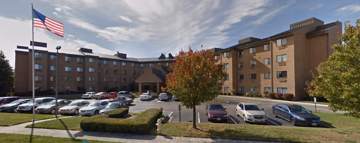 Exterior view of a multi-story brick building with multiple windows, a parking lot with several parked cars, an American flag on a flagpole, and trees with autumn foliage in front of the building under a partly cloudy sky.