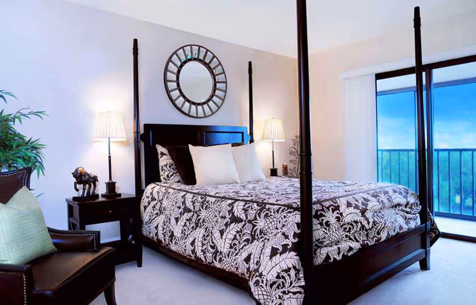 Bright bedroom with a dark four-poster bed dressed in patterned bedding, two bedside lamps, an armchair, and a sliding glass door leading to a balcony.