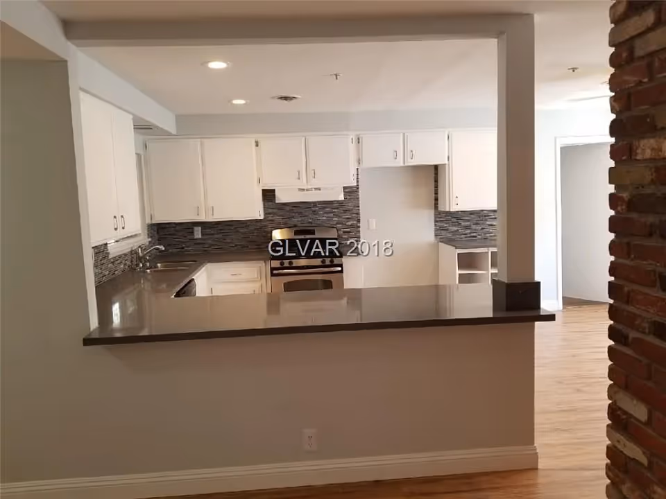 View of a modern kitchen with white cabinets, a stainless steel stove, a tiled backsplash, and a countertop bar area. The kitchen opens into a room with wooden flooring and a brick column on the right side.