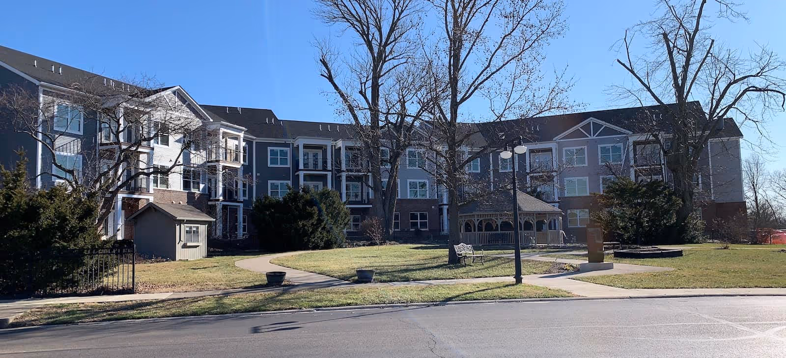 Exterior view of a multi-story senior living facility building with a well-maintained lawn, leafless trees, a gazebo, benches, and a paved walkway under a clear blue sky.