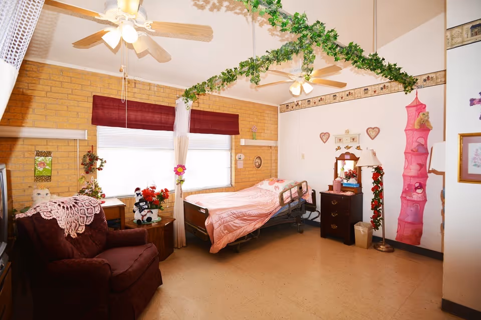 A decorated nursing-home bedroom with a single bed dressed in pink, an armchair, a dresser, ceiling fans, and hanging greenery.
