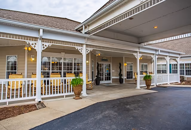 Covered entrance area of a senior living facility with white railings, potted plants, and yellow cushioned chairs on a porch. The building has beige siding and multiple windows, with a welcome sign above the glass double doors.
