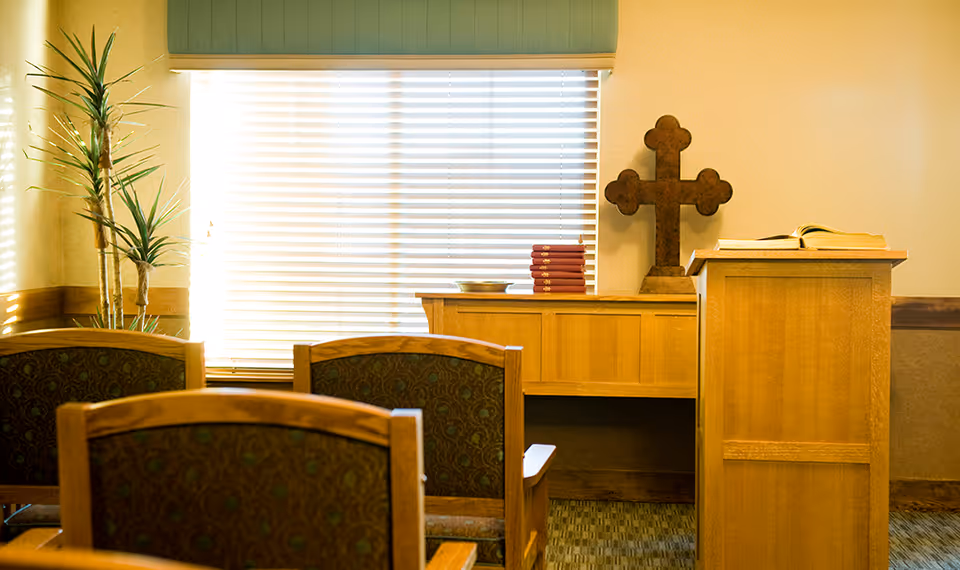 Small chapel-style interior with wooden chairs, a lectern, a cross on a side table, and window blinds.