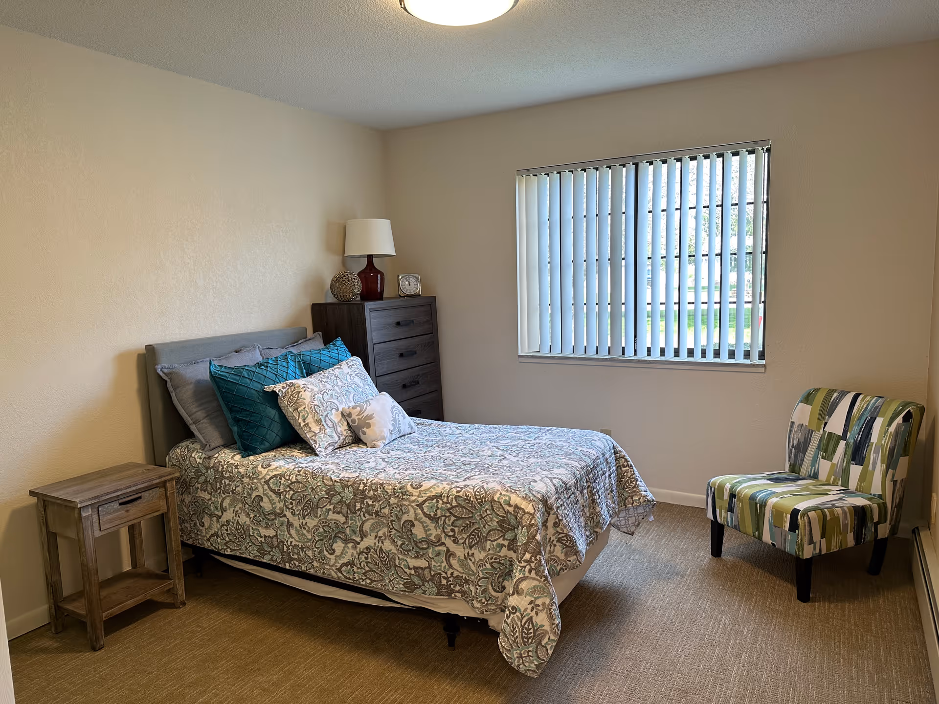 A simple and cozy bedroom with a bed covered in a patterned quilt and multiple pillows. Next to the bed is a wooden nightstand and a tall dresser with a lamp and decorative items on top. A window with vertical blinds lets in natural light, and a colorful upholstered chair is placed near the window.