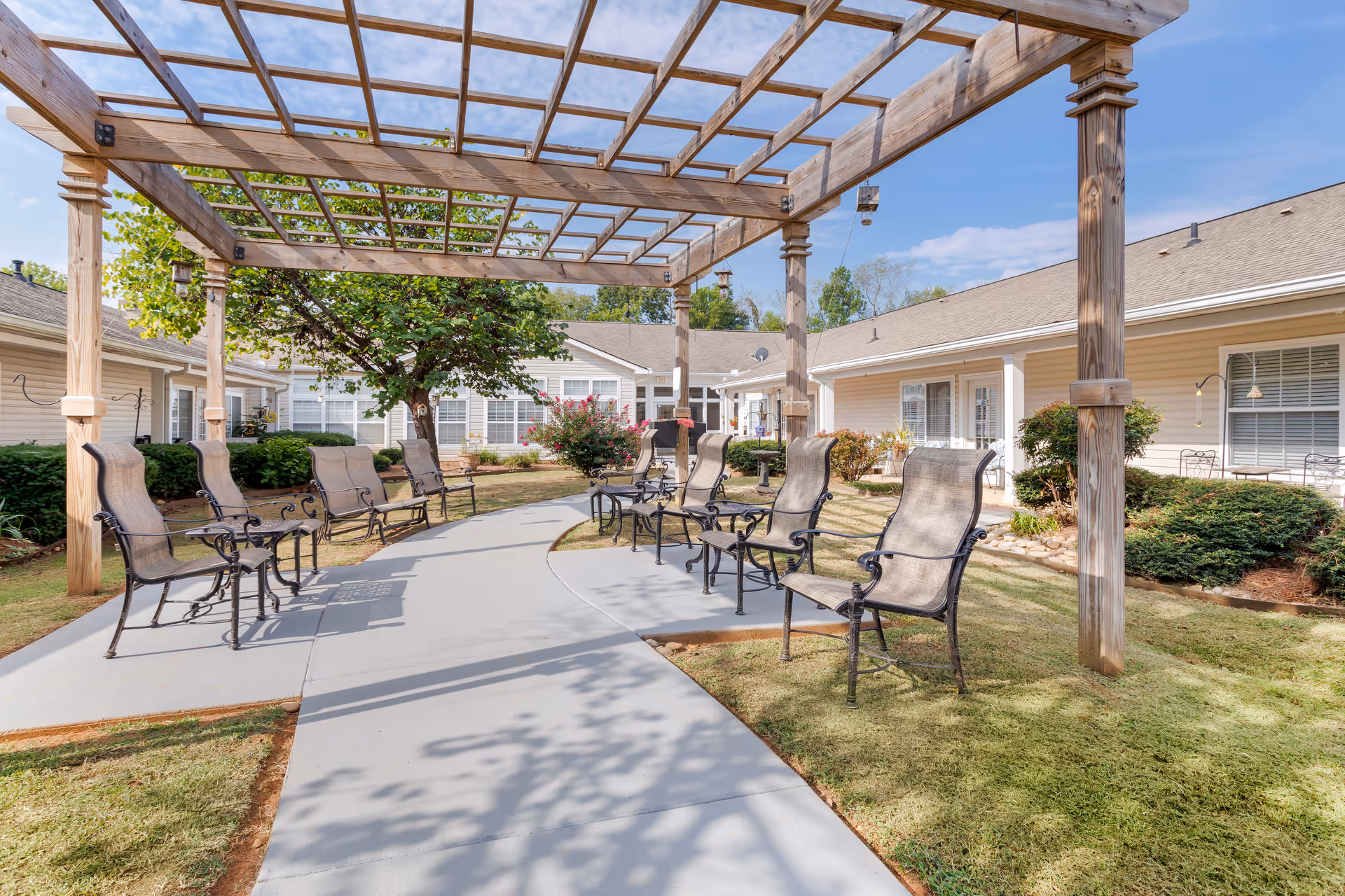 Outdoor seating area with multiple metal chairs arranged under a wooden pergola in a courtyard surrounded by single-story buildings with beige siding and white trim. The area has a concrete pathway, green grass, bushes, and a tree with a clear blue sky above.