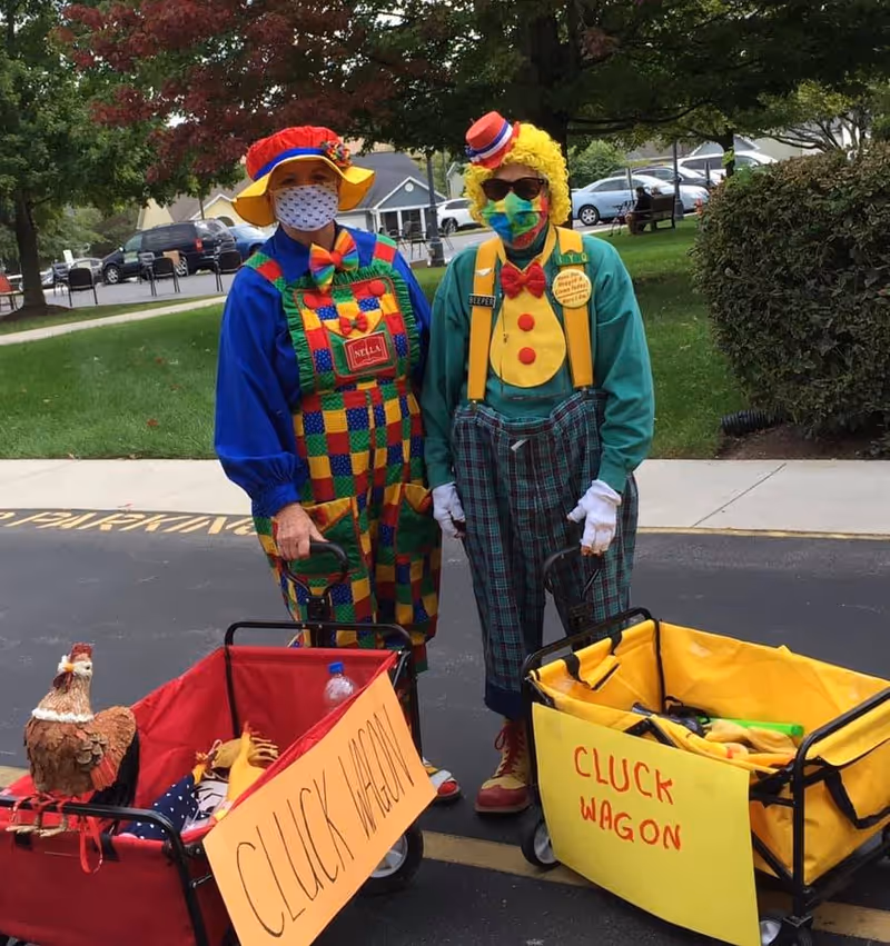 Two people dressed as clowns wearing colorful costumes and face masks standing outdoors on a paved area with grass and trees in the background. Each person is holding a small wagon labeled 'CLUCK WAGON,' one red and one yellow. There is a decorative chicken figure in the red wagon.