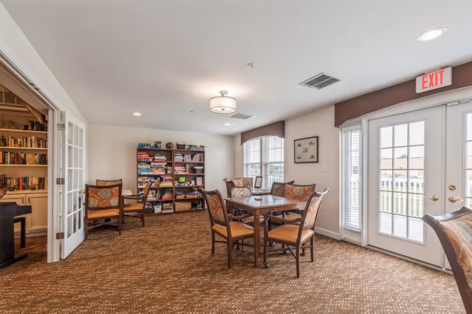Sunlit common room with a table and chairs, book and game-filled shelves, and French doors leading outside.