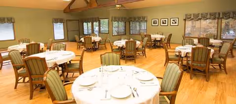 Dining room in a senior care center with round tables set for meals and wooden chairs.