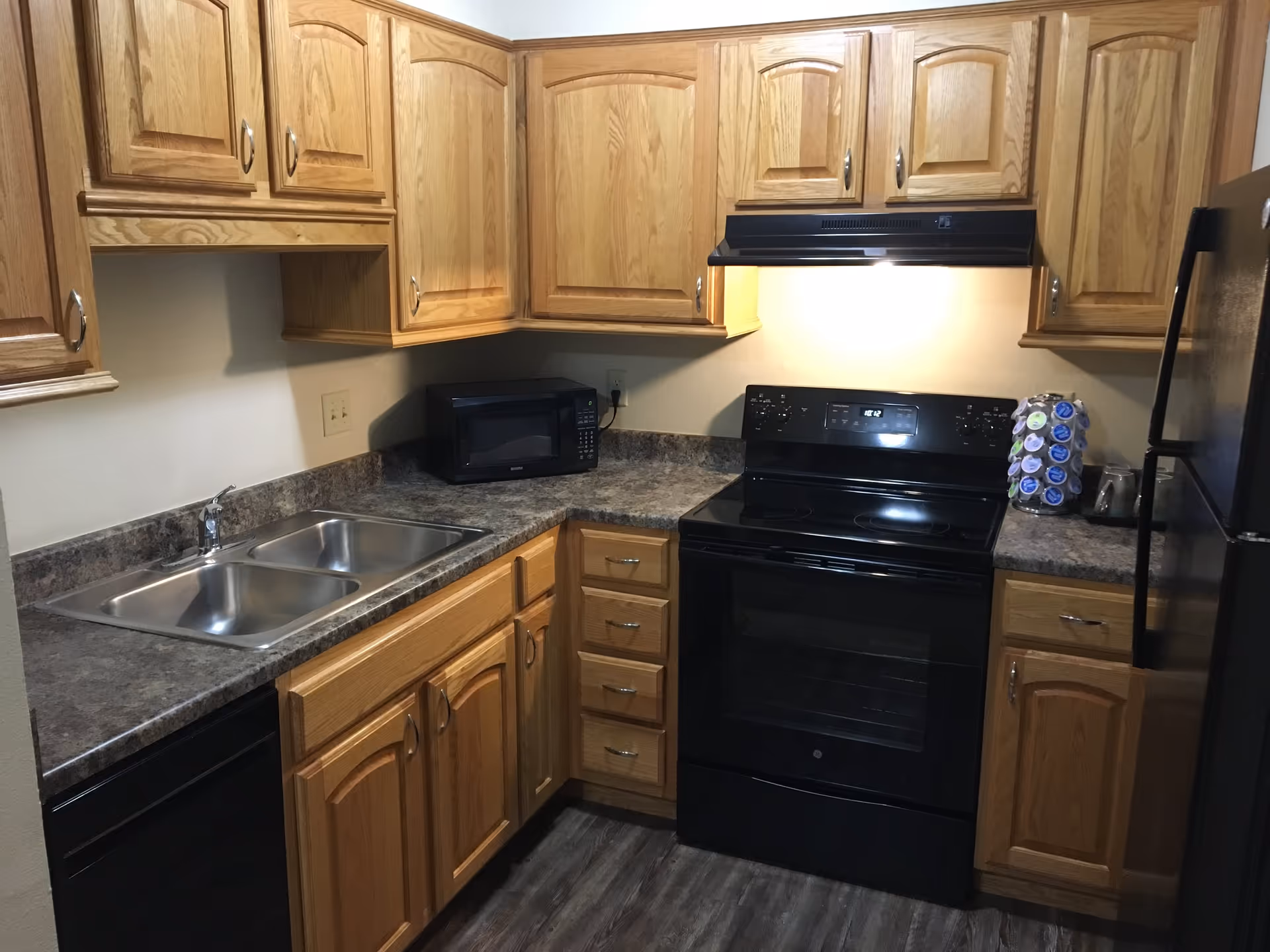 A compact kitchen with wooden cabinets, a double stainless steel sink, a black microwave, a black electric stove with an overhead vent, and a black refrigerator. The countertop is a dark marbled pattern, and there is a rotating holder with coffee pods next to the stove. The floor has a wood-like finish.
