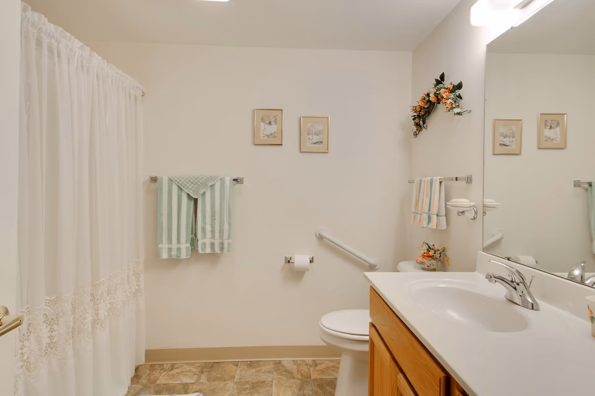 Bathroom with a sink vanity, toilet with a grab bar, towel racks holding striped towels, decorative wall art, and a shower with a lace curtain.