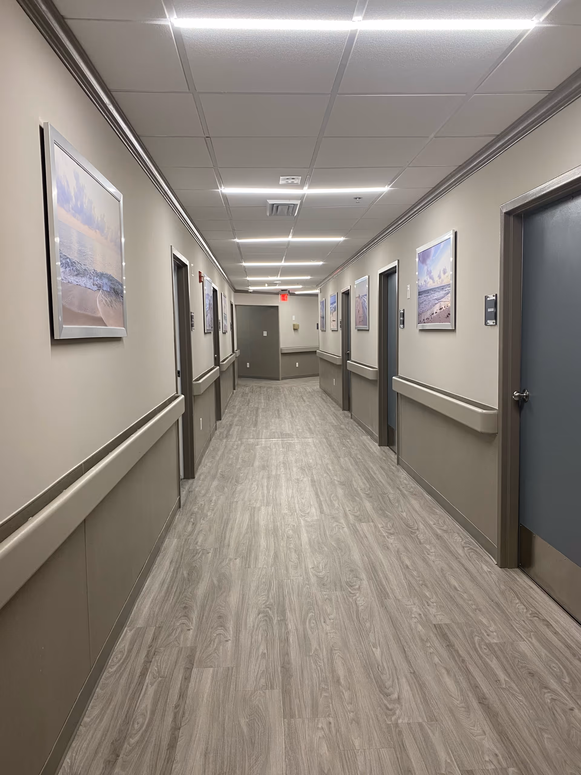 A long, well-lit hallway in a senior living facility with light wood flooring, beige walls, and several closed doors on both sides. The walls are decorated with framed pictures of beach scenes, and there are handrails along both sides of the hallway.
