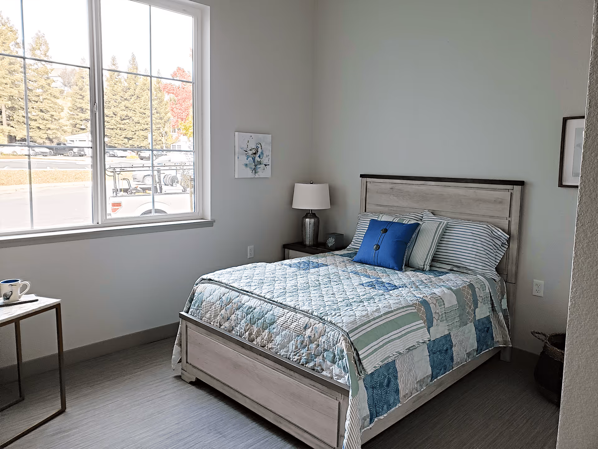 A bright bedroom with a large window showing an outdoor view of trees and parked cars. The room features a wooden bed with a patchwork quilt in shades of blue, green, and white, two pillows including a blue decorative one, a bedside table with a lamp, a small side table with a coffee cup, and minimal wall art.