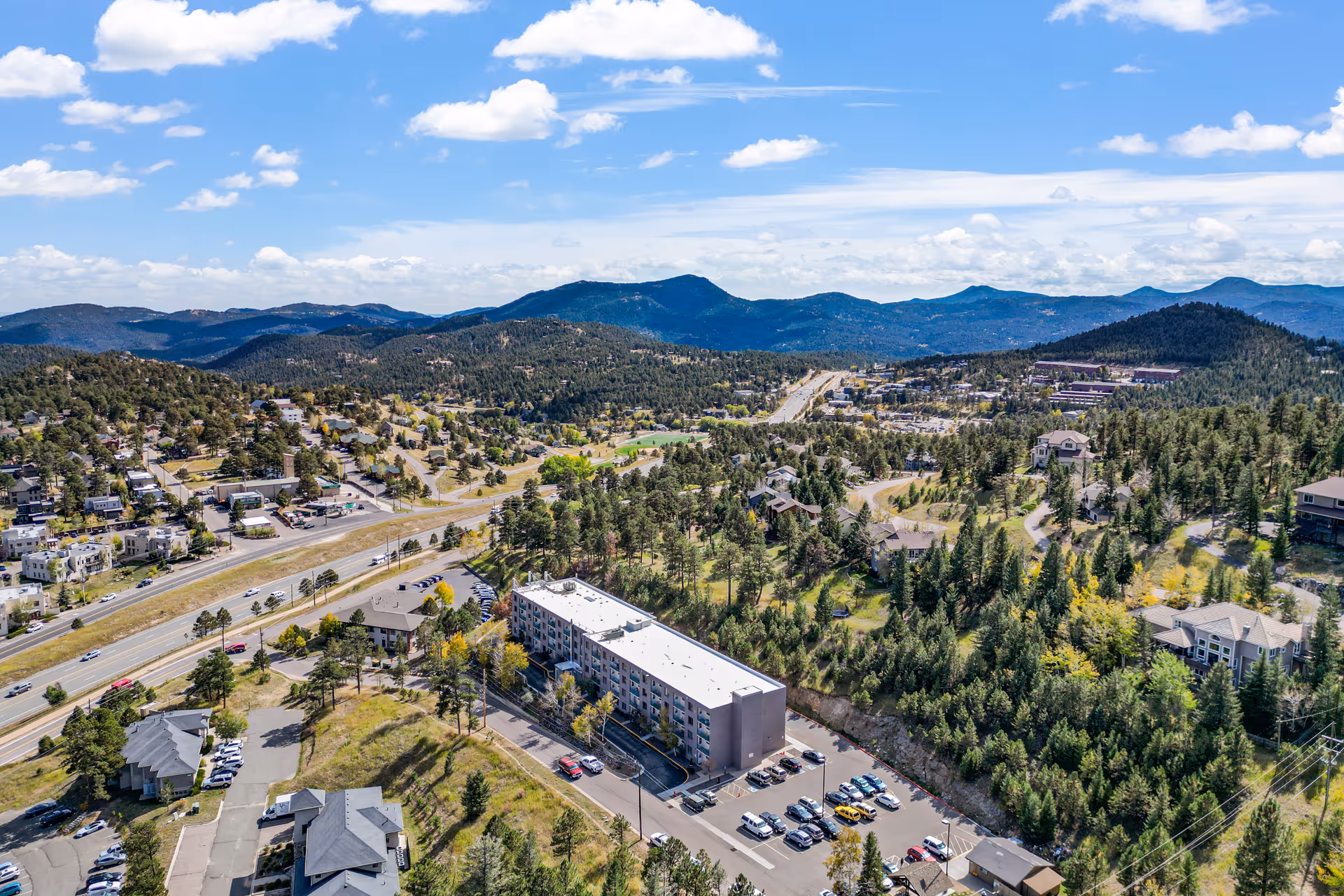 Aerial view of a mountainous area with a large building surrounded by trees and parking lots, roads, and other smaller buildings scattered throughout the landscape under a partly cloudy blue sky.