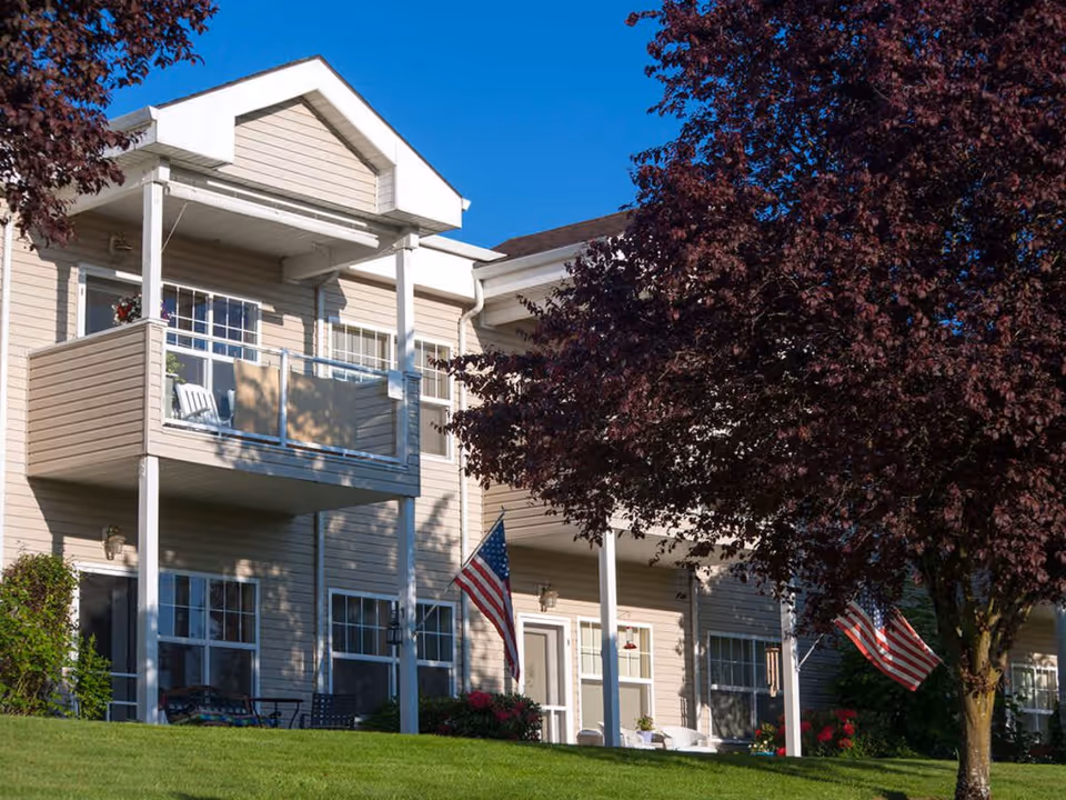 Exterior view of a beige two-story senior living building with balconies, American flags, and a tree in front.