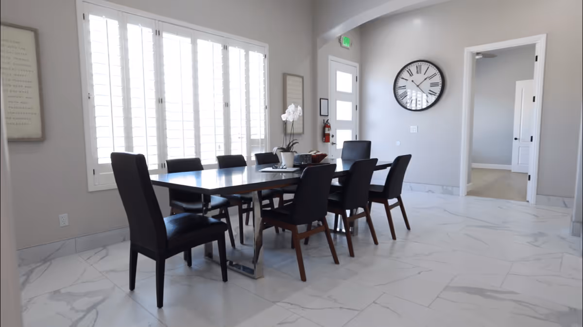 A modern dining room with a long rectangular table surrounded by eight black chairs. The room has large windows with white shutters allowing natural light to fill the space. There is a large wall clock above a doorway, a fire extinguisher mounted on the wall near the door, and light-colored marble flooring.