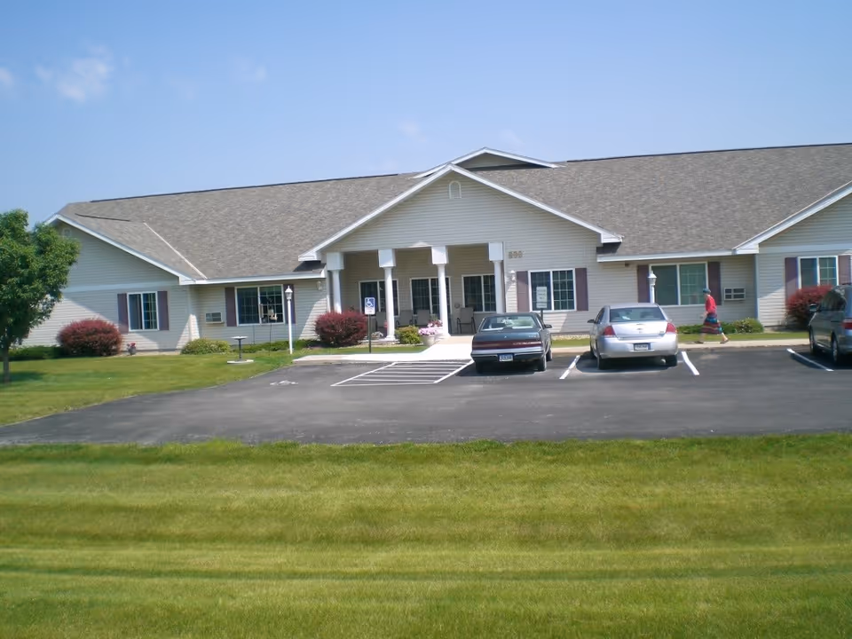Single-story building with beige siding and a gray shingled roof, featuring a covered entrance supported by white columns. There are three cars parked in front, a person walking on the sidewalk, and well-maintained green grass and bushes surrounding the building under a clear blue sky.