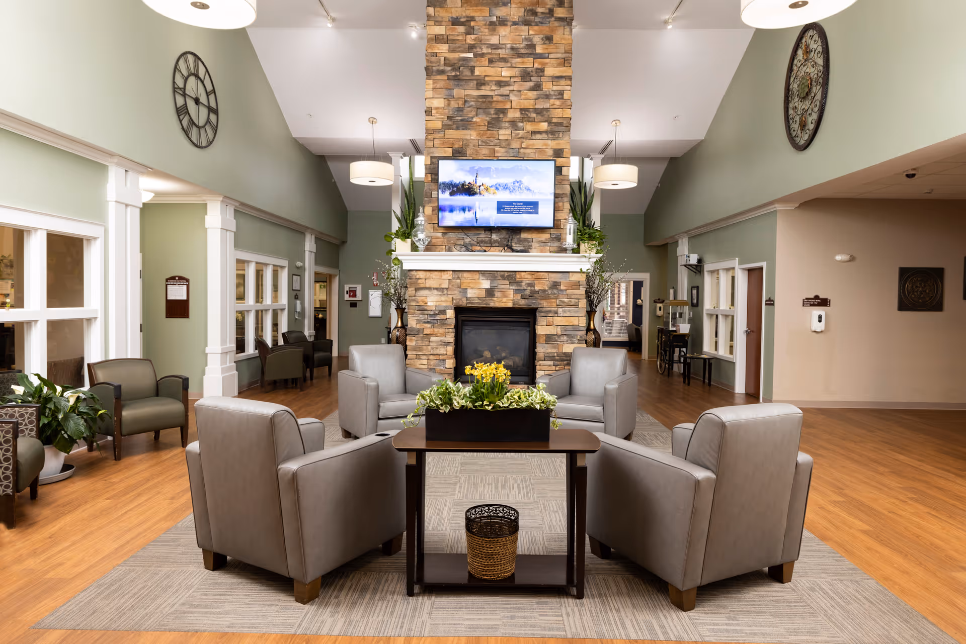 A spacious and well-lit senior living facility common area with a stone fireplace in the center. Four gray armchairs are arranged around a wooden coffee table with a planter of yellow flowers. The room has light green walls, wooden flooring, large wall clocks on either side, and a flat-screen TV mounted above the fireplace. Additional seating and plants are visible along the walls.