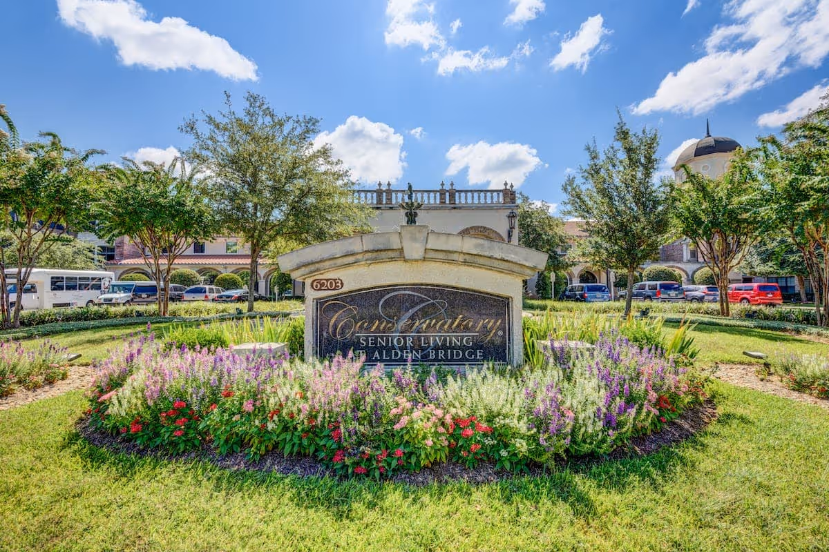 Outdoor view of the Conservatory Senior Living at Alden Bridge facility sign surrounded by colorful flowers and greenery, with the building and parked cars visible in the background under a partly cloudy blue sky.