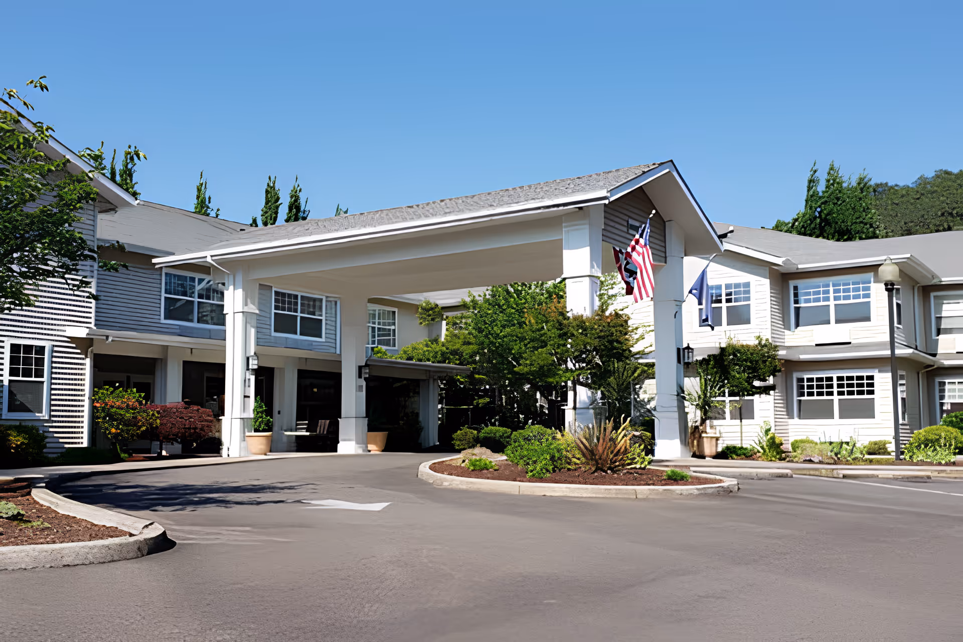 Exterior view of Oak Park Senior Living by Cogir showing a large covered entrance with white pillars, an American flag, and well-maintained landscaping including bushes and trees under a clear blue sky.