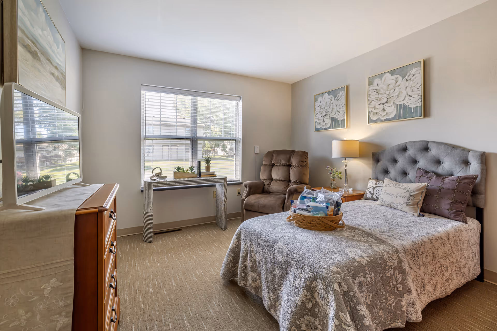 A cozy bedroom in a senior living facility featuring a bed with a gray floral quilt and decorative pillows, a brown recliner chair, a wooden nightstand with a lamp and flowers, a window with blinds letting in natural light, a dresser with a TV on top, and two floral paintings on the wall.