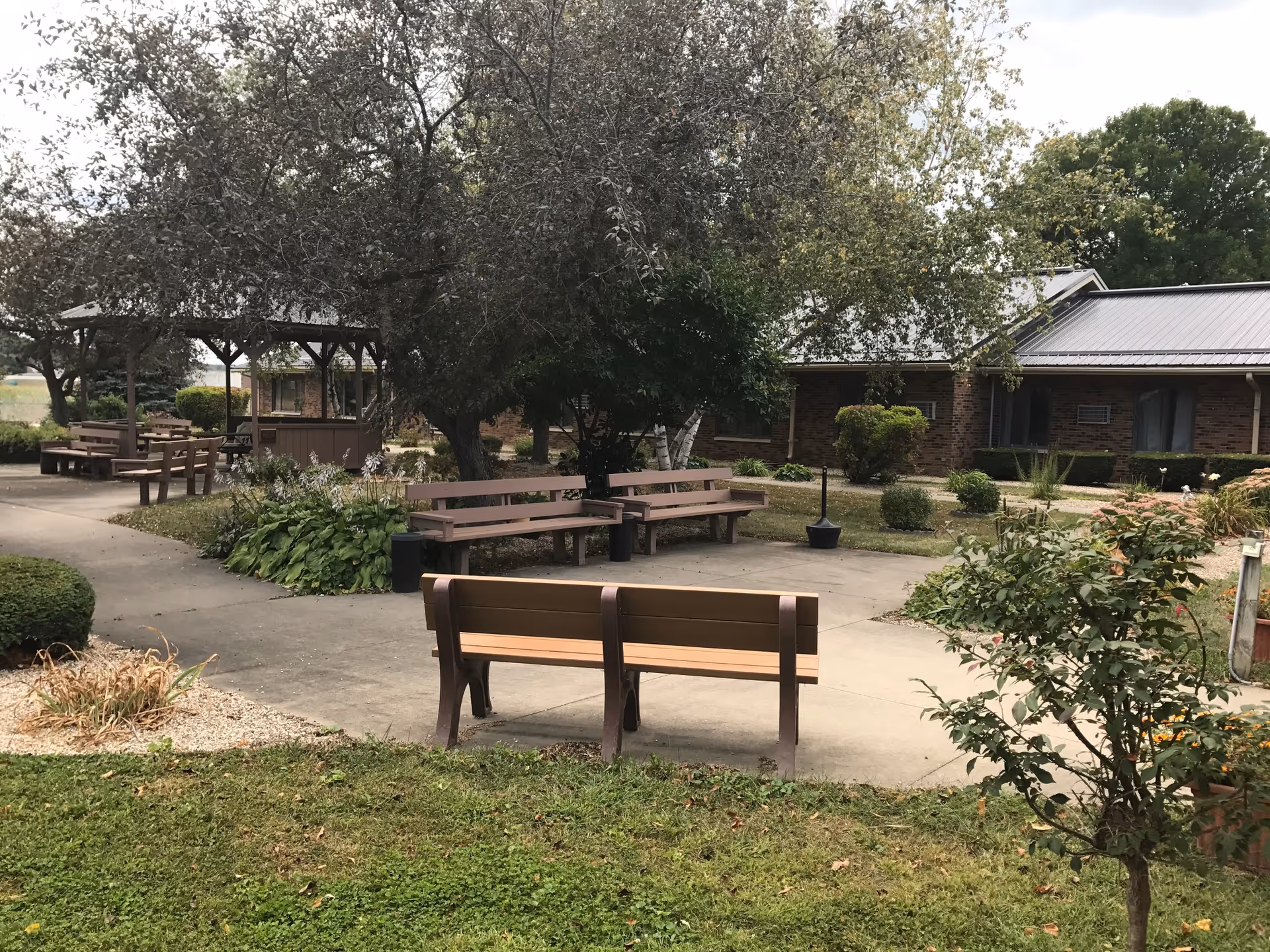 Outdoor seating area in a garden with several wooden benches and picnic tables under trees and a gazebo, surrounded by shrubs and plants, with a brick building in the background.