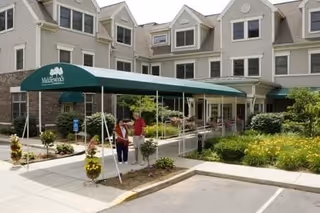 Entrance of Masonicare at Middlewoods of Farmington facility with a green canopy over the walkway. Two people are standing near the entrance, surrounded by landscaped plants and flowers. The building is a multi-story structure with beige siding and multiple windows.