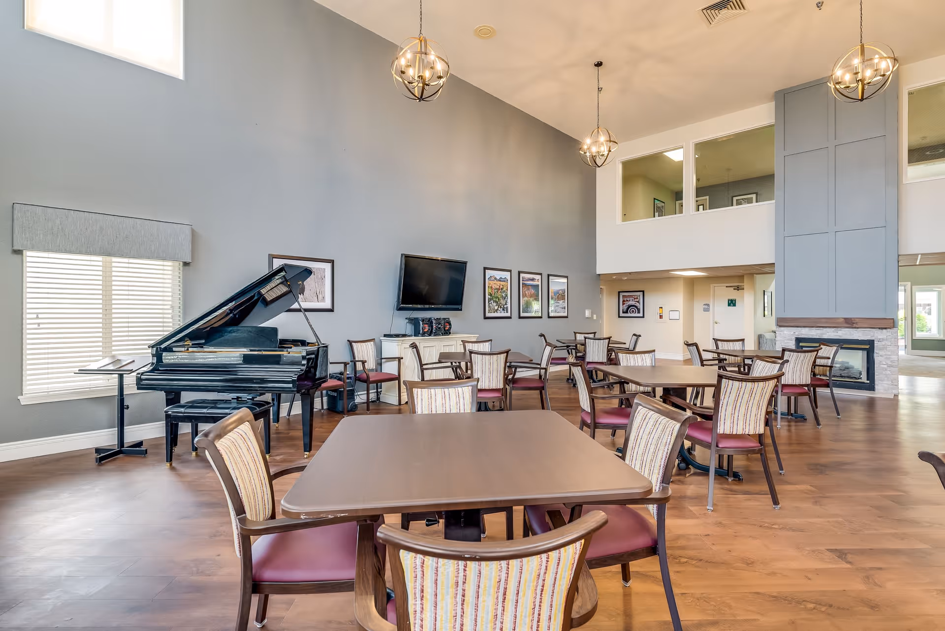 A spacious senior living common area with multiple tables and chairs arranged for socializing or dining. There is a black grand piano near a window with blinds, a wall-mounted TV, and framed pictures on the gray wall. The room features high ceilings with modern hanging light fixtures and a fireplace with a stone surround.