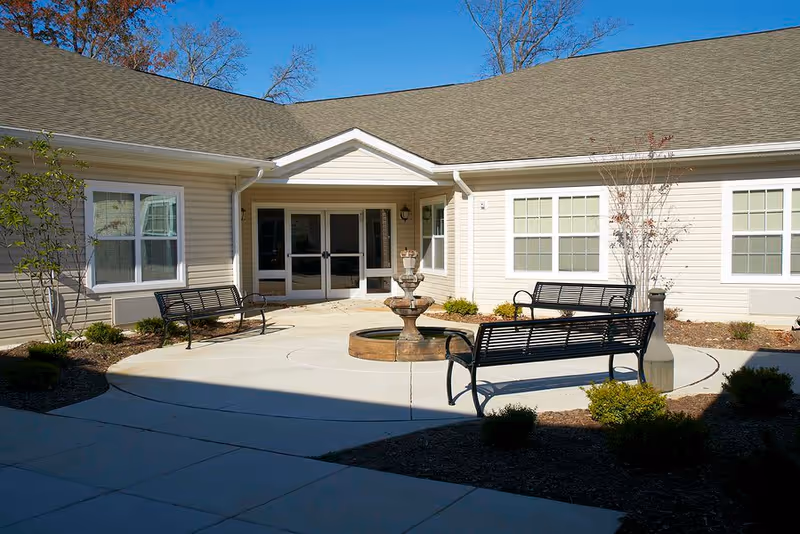 Outdoor courtyard area of a senior living facility with a central water fountain surrounded by three black metal benches, small bushes, and a beige building with multiple windows and a double glass door entrance under a clear blue sky.