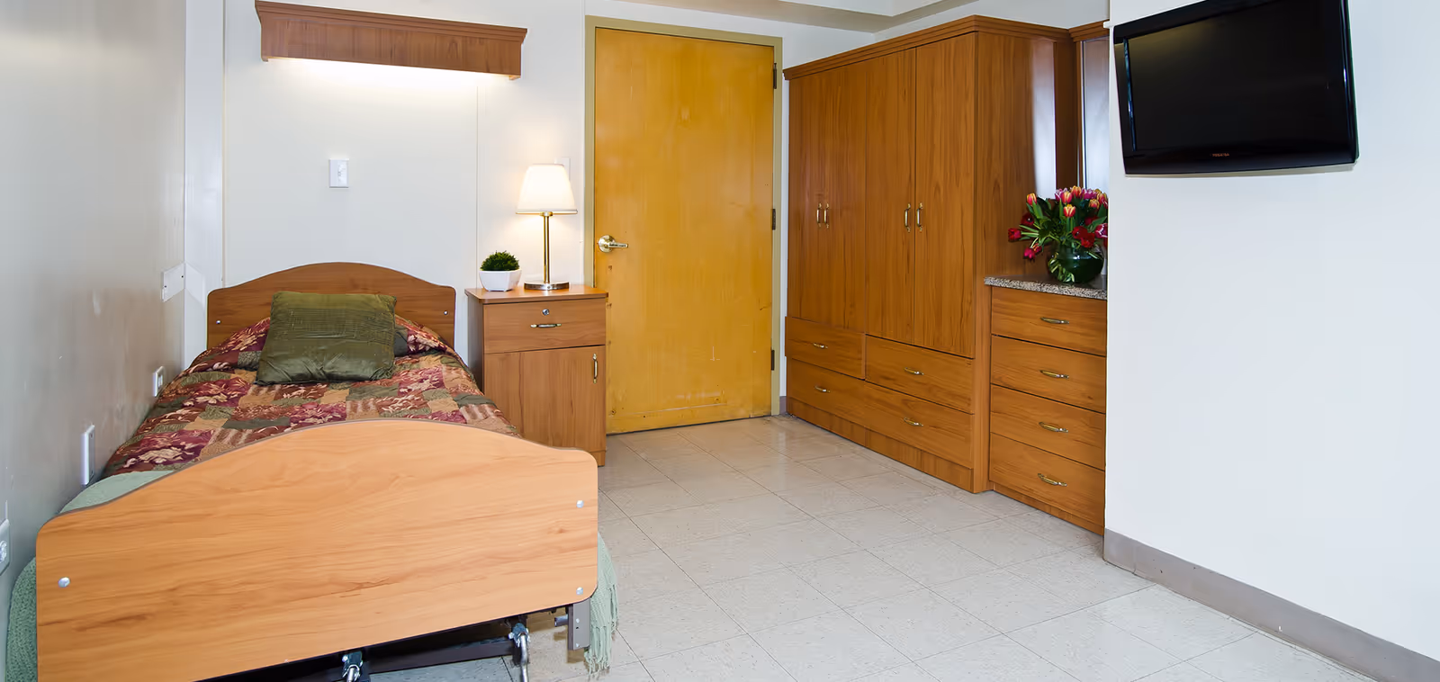 A small bedroom with a single wooden bed covered with a patterned quilt and a green pillow. Next to the bed is a wooden nightstand with a lamp and a small plant. Across from the bed is a wooden wardrobe and dresser with a vase of flowers on top. A flat-screen TV is mounted on the wall to the right. The room has a tiled floor and a wooden door.