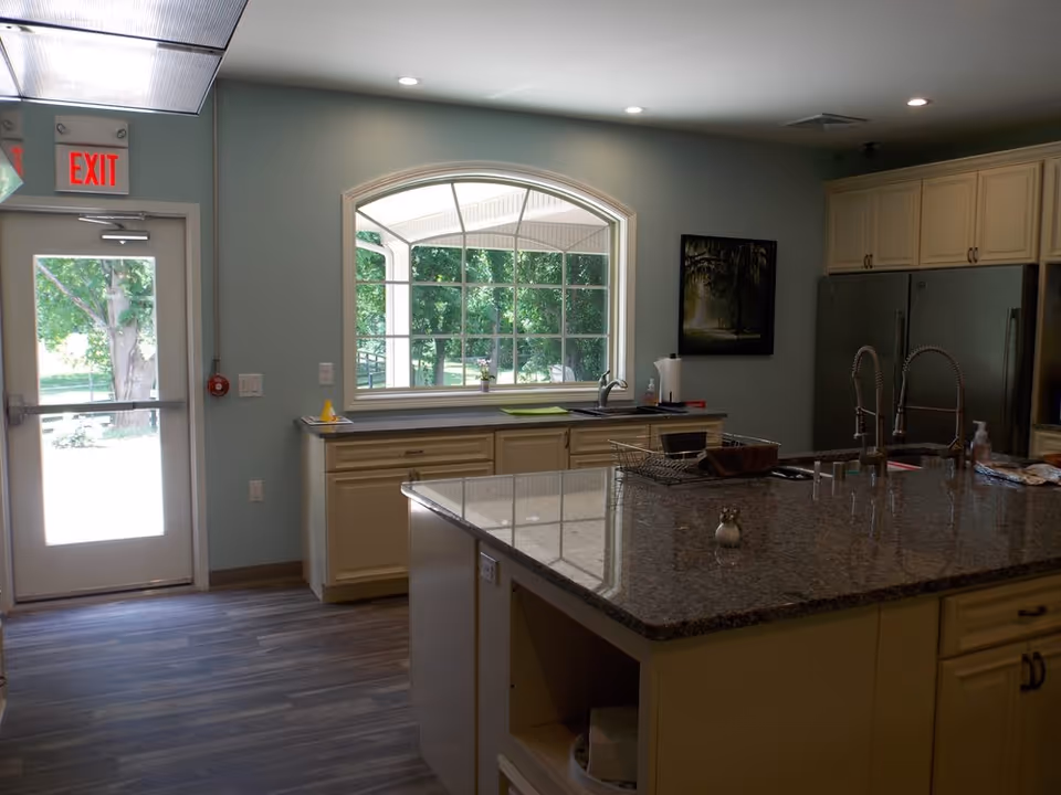 Interior view of a kitchen in Pleasant Meadow Assisted Living featuring a large granite island countertop with two modern faucets, cream-colored cabinets, a window with a view of green trees outside, a double-door refrigerator, and an exit door with an illuminated red exit sign above it.