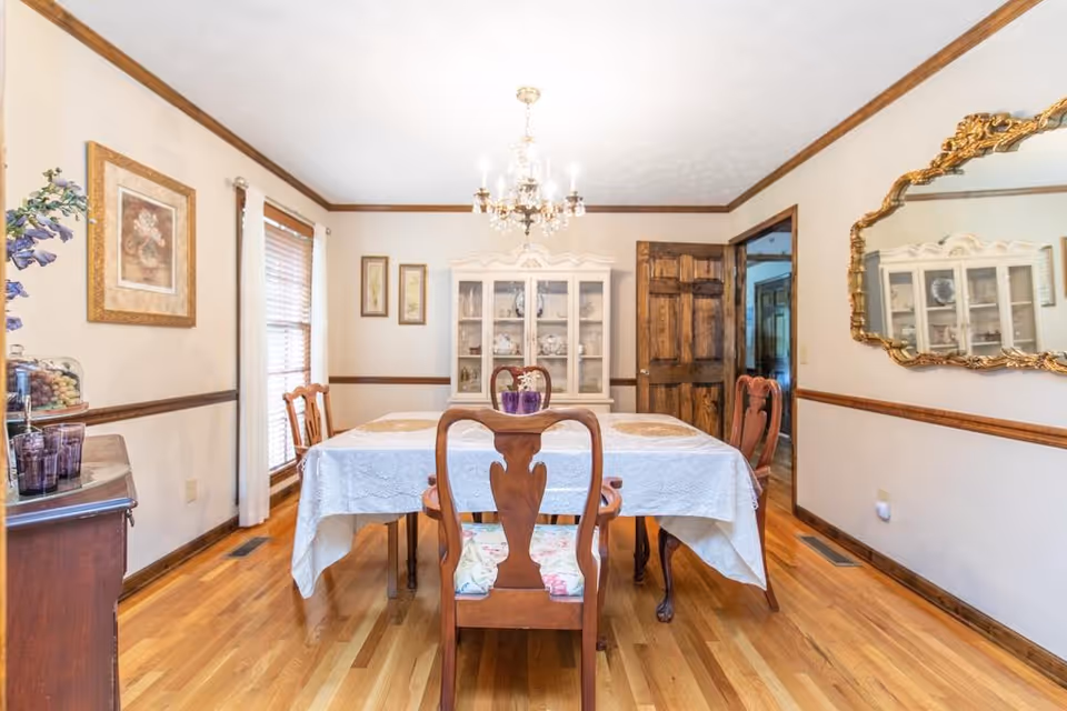 Formal dining room with a table covered by a white tablecloth, wooden chairs, a chandelier, china cabinet, and an ornate wall mirror.