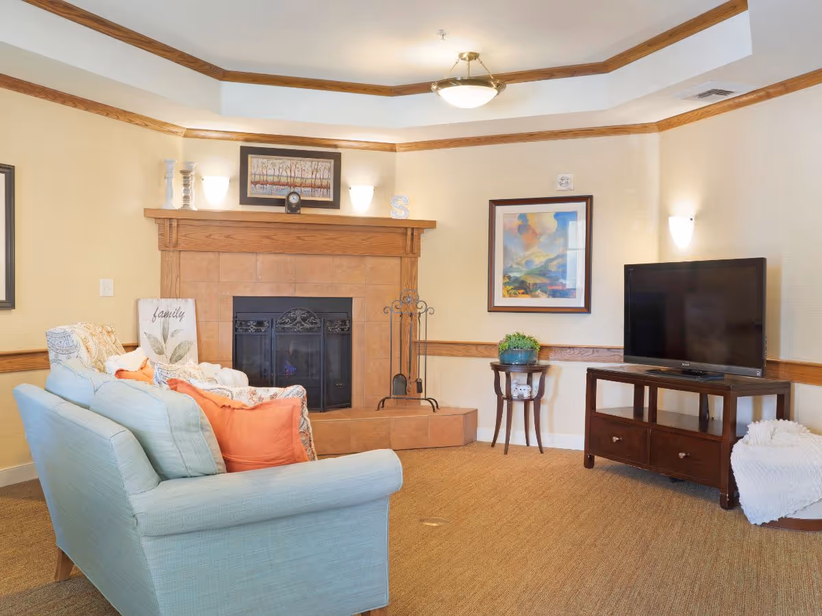 A cozy living room with a light blue couch adorned with orange and patterned pillows facing a fireplace with a wooden mantle. Above the mantle is a framed painting and two wall sconces. To the right, there is a flat-screen TV on a wooden stand with a small round table holding a plant beside it. The room has beige walls with wooden trim and a carpeted floor.