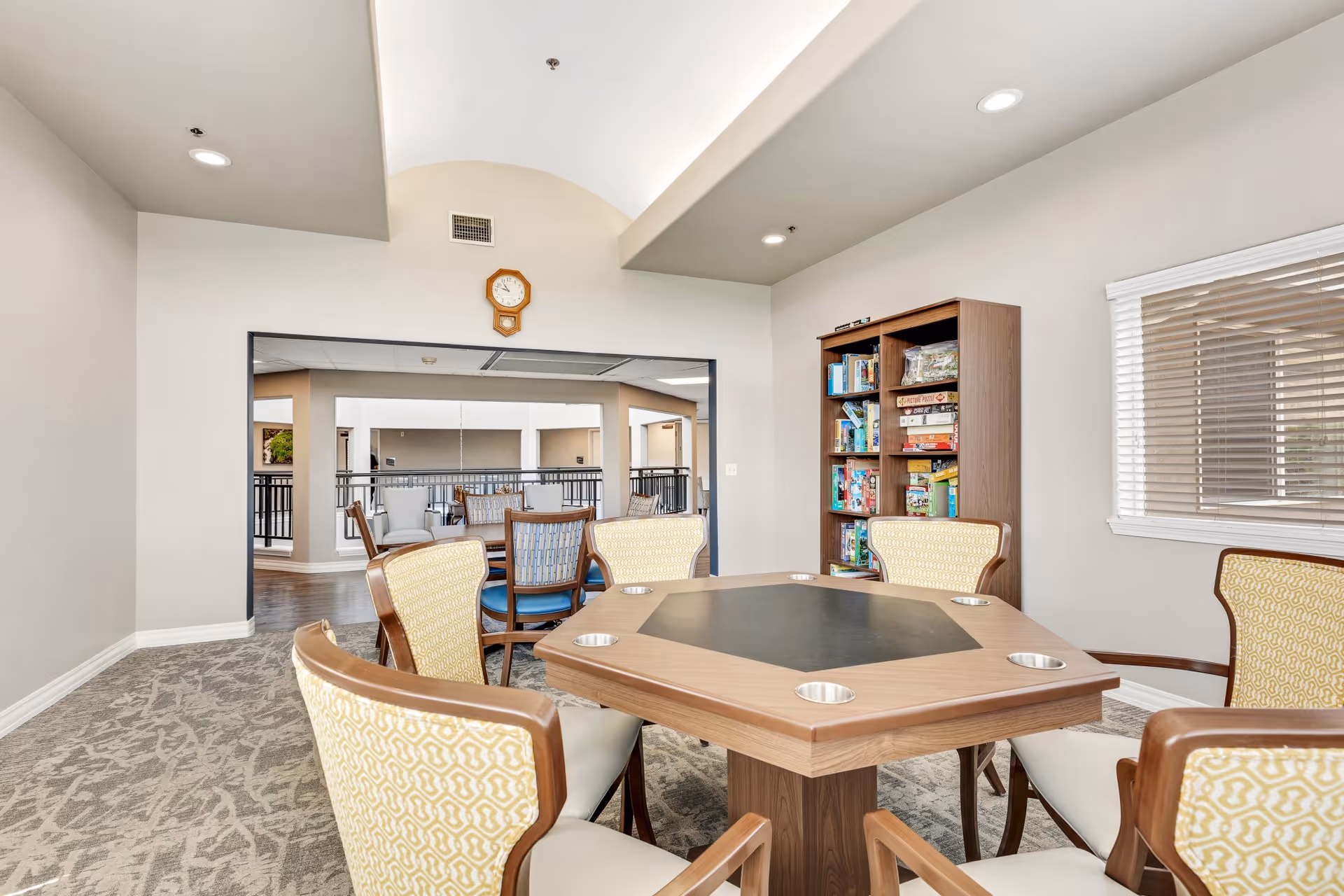A bright and spacious common area with a hexagonal wooden table surrounded by six cushioned chairs with yellow patterned backs. Behind the table is a bookshelf filled with board games and puzzles. The room has a large window with blinds and a wall clock above a wide opening that looks into another seating area with additional chairs and tables.