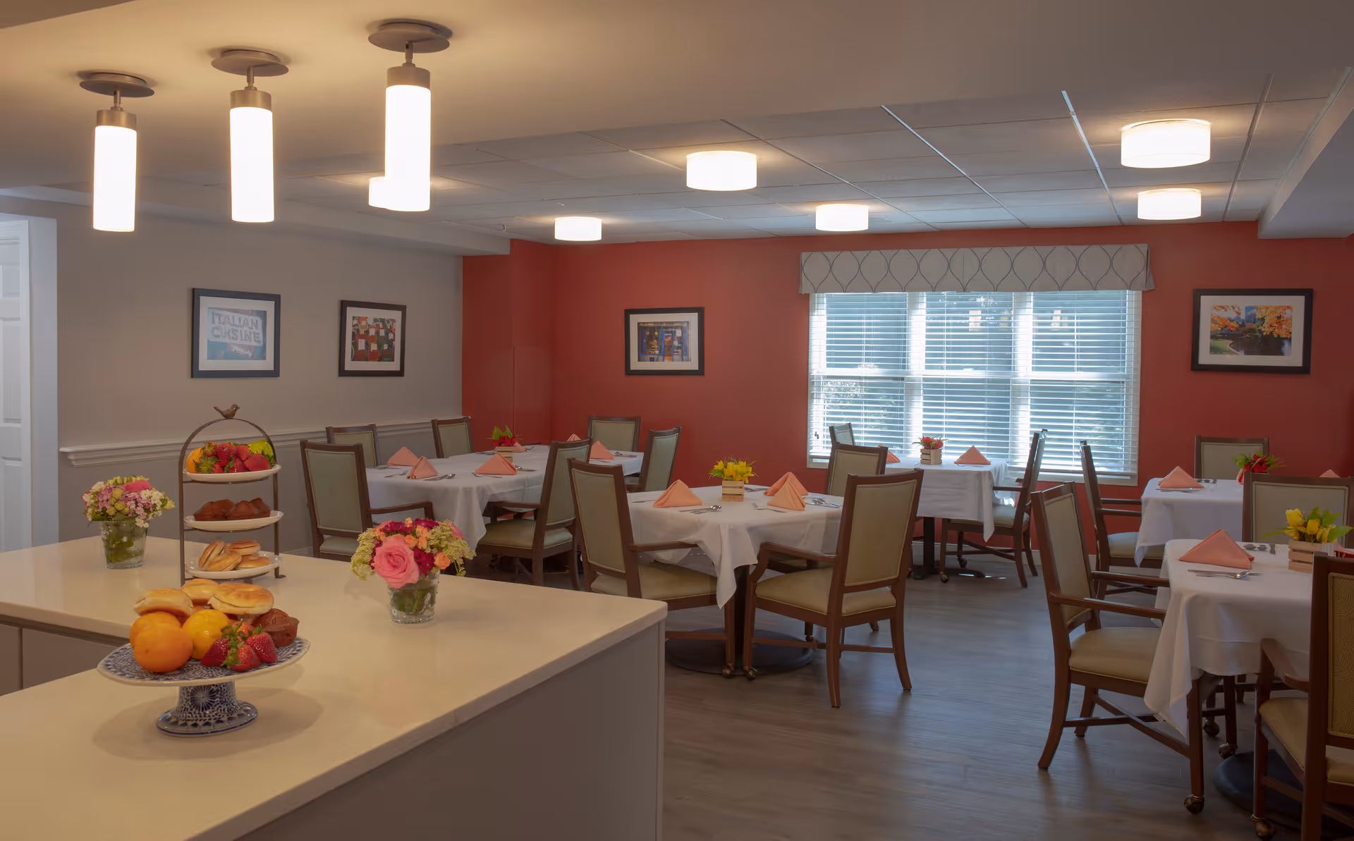 A dining room in an assisted living facility with several tables covered in white tablecloths, each set with folded pink napkins and small flower arrangements. The room has a red accent wall with framed artwork and a large window with blinds. In the foreground, a counter displays plates of muffins, bagels, fruit, and small flower vases. Ceiling lights illuminate the space warmly.
