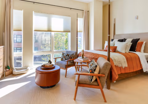 Sunlit bedroom with a four-post bed dressed in rust-orange bedding, two mid-century chairs and an ottoman beside large windows.