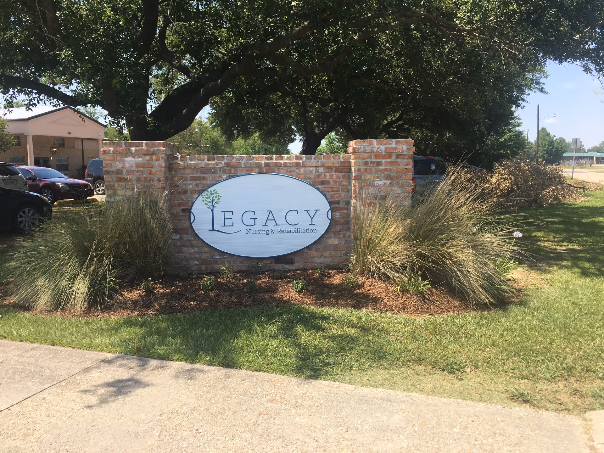 Outdoor view of a brick sign for Legacy Nursing & Rehabilitation surrounded by grass, plants, and trees with parked cars and a building in the background.