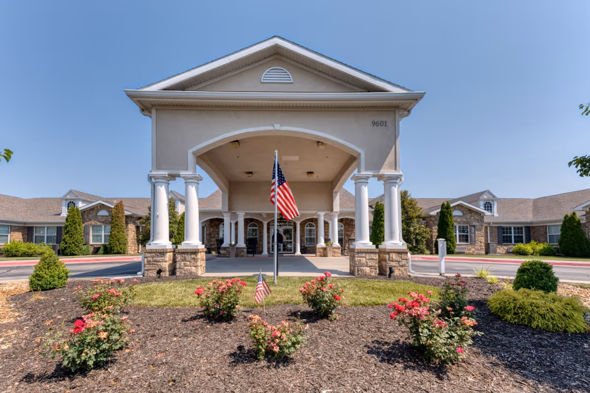 Front exterior view of Addington Place of Shoal Creek building with a covered entrance supported by white columns, an American flag in front, and landscaped flower beds with red flowers and green shrubs under a clear blue sky.